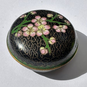 Könnte beinhalten: Eine kleine, runde, schwarze Cloisonné-Box mit Deckel. Die Box ist mit einem floralen Design aus rosa und weißen Blüten mit grünen Stielen und Blättern verziert. Der Hintergrund ist ein schwarzes und goldenes Muster.