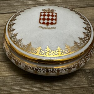 May include: A white porcelain trinket box with a gold trim and a red and white checkered coat of arms with a crown on top. The box says "MONACO" in gold lettering.