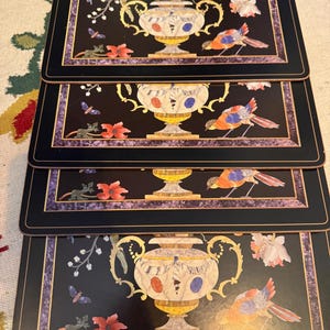 May include: Set of four black placemats with a decorative design. Each placemat features a central vase with colourful flowers, birds, and butterflies. The border is a dark purple with gold accents.