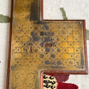 May include: An antique brass and wood decorative piece. The brass has an ornate, engraved design with a keyhole and a grid pattern. The wood frame is a dark brown color. The piece is likely from a cabinet or door.