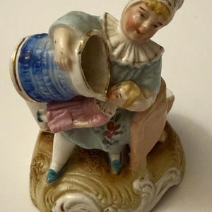 May include: A porcelain figurine depicting a person pouring from a blue and gold pitcher. The figure wears a light blue dress with floral accents, a white ruffled collar, and a white cap with red floral details. The figurine is set on a decorative base.