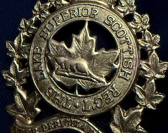 May include: A gold-colored metal badge with a crown on top and a maple leaf with a beaver inside. The text around the maple leaf reads "LAKE SUPERIOR SCOTTISH REG'T."