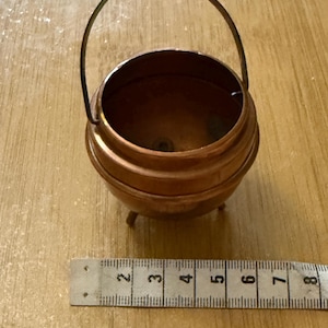 May include: A small, copper-coloured cauldron with a curved handle and three small feet. The cauldron is approximately 7.6 cm in diameter. A ruler is placed next to the cauldron for scale.
