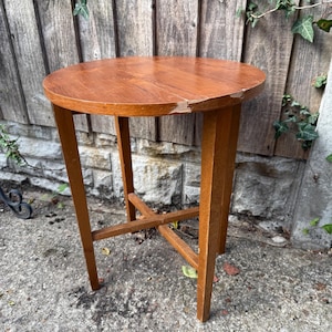 May include: A small, round wooden table with a warm brown finish. The table has a three-legged design with an X-shaped support near the base. The tabletop shows some wear. The table is approximately 61 cm tall.