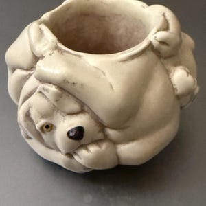 May include: A white ceramic planter shaped like a dog with its head tucked into its paws. The planter has a small opening at the top for planting.