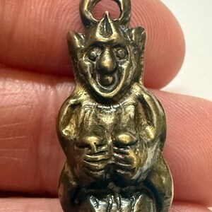 May include: A small, antique-style brass figurine of a smiling, humanoid creature with large ears and a crown-like headpiece. The figure has its hands clasped in front of its chest and a loop at the top for hanging. The overall color is a tarnished gold.