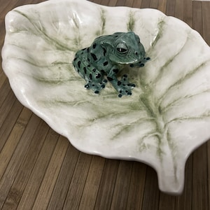Puede incluir: Un plato de cerámica blanca con forma de hoja con detalles pintados en verde, con una figurita de rana verde y negra sentada en la hoja.