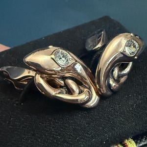 May include: A pair of metallic silver-toned cufflinks shaped like coiled snakes. Each snake head features a single clear, faceted crystal set into the top. The metal has a polished, reflective finish, and the design shows the snake body looping.