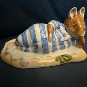 以下が含まれることがあります： 青い白のストライプの寝巻きを着たネズミの陶器の置物。ビーチタオルの上に横たわっています。ネズミは貝殻と海藻のある砂のような土台の上にあります。