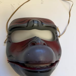 May include: A vintage Japanese Noh theater mask. The mask features a dark red and black color scheme with a prominent nose and open mouth revealing teeth. A brown strap is attached for wearing.