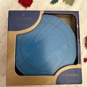 May include: A set of six blue coasters with a wood grain design, packaged in a square box. The box has gold trim and the brand name "Pimpernel" is printed on the front. The coasters are round with a gold ring design.