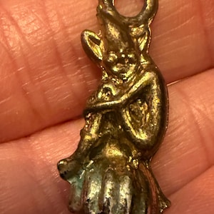 May include: A small, antique-looking charm in the shape of a seated figure, possibly a gnome or elf. The charm is made of a gold-toned metal with a loop at the top for hanging. The figure is detailed with visible facial features and clothing.