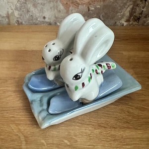 May include: Two white ceramic bunny salt and pepper shakers on a blue and white ceramic tray. The bunnies are wearing colourful scarves and are skiing on blue skis.