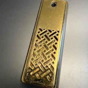 Könnte beinhalten: Eine rechteckige, goldfarbene Parfümflasche mit einem dekorativen geometrischen Muster auf der unteren Hälfte. Die Flasche hat oben ein kleines Loch und das Wort "Guerlain" ist unten aufgedruckt.