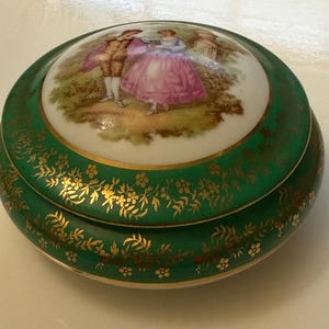 Könnte beinhalten: Eine antike Porzellan-Schmuckdose mit grünem Sockel und goldenen Verzierungen. Der Deckel zeigt eine bemalte Szene eines Paares in einem Garten mit dem Wort "promenade" in Gold.