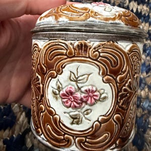 Può includere: Un barattolo in ceramica vintage con coperchio in metallo. Il barattolo cilindrico presenta un motivo floreale marrone e bianco. Il coperchio ha un piccolo pomello decorativo. Il barattolo è tenuto in una mano.