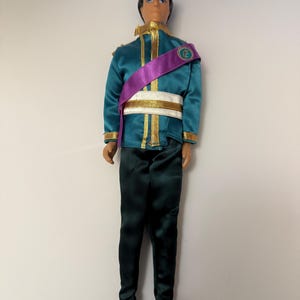 Puede incluir: Mu&ntilde;eco vestido con una chaqueta verde azulado con ribetes dorados, una faja morada y pantalones verde oscuro. El mu&ntilde;eco tiene pelo negro y lleva zapatos negros. La chaqueta tiene detalles dorados y un cintur&oacute;n blanco.