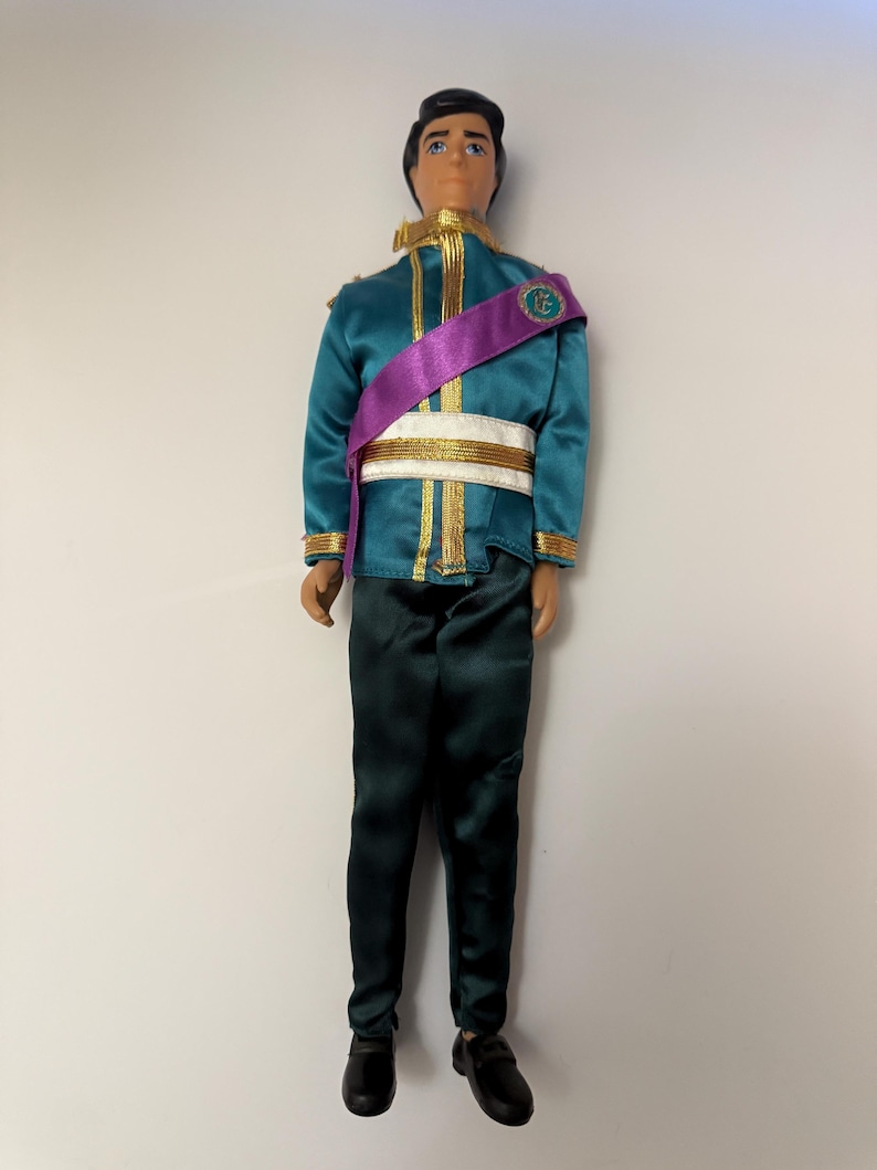 Puede incluir: Mu&ntilde;eco vestido con una chaqueta verde azulado con ribetes dorados, una faja morada y pantalones verde oscuro. El mu&ntilde;eco tiene pelo negro y lleva zapatos negros. La chaqueta tiene detalles dorados y un cintur&oacute;n blanco.