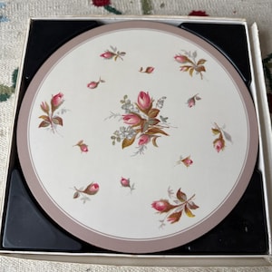 May include: A set of round coasters featuring a floral rose pattern. The coasters have a white centre with pink roses and green leaves, framed by a pink border. The coasters are presented in a black and white box.