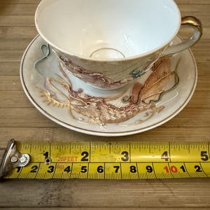 May include: A white teacup and saucer with a gold rim and a dragon design. The dragon is depicted in shades of brown, gold, and pink. The saucer has a gold rim and a dragon design in shades of brown, gold, and pink.