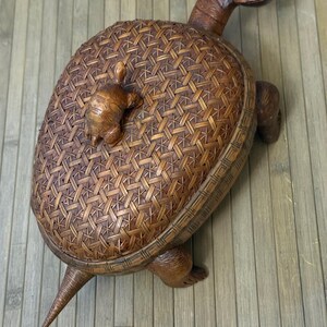 May include: A handcrafted wooden turtle-shaped basket with a woven pattern on its shell. A smaller turtle figure is attached to the top. The turtle has a long neck, a pointed tail, and four legs. The basket is brown.