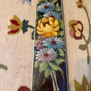 Op de afbeelding: Een rechthoekig, handbeschilderd decoratief item met een boeket bloemen. De bloemen omvatten een grote gele bloem, blauwe en witte madeliefjes en kleinere roze bloemen, alles tegen een donkerblauwe achtergrond. Het item heeft een witte rand.