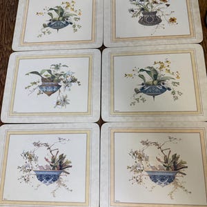 May include: A set of six rectangular placemats featuring a cream-colored background with a light tan border. Each placemat displays a central illustration of a decorative blue and white patterned bowl filled with various floral arrangements.