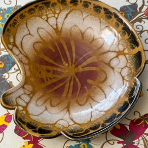 Puede incluir: Un plato de cerámica con un diseño marrón, blanco y dorado. El plato tiene un borde festoneado y un patrón floral.