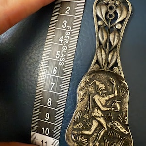 May include: A silver-toned metal spoon, approximately 10 inches long, with an ornate design. The handle features a floral motif, while the bowl depicts a figure in a forest scene. The spoon is held next to a measuring tape.