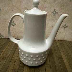 Puede incluir: Cafetera de cerámica blanca con tapa abovedada y base texturizada. La cafetera tiene un pico redondeado y un asa.