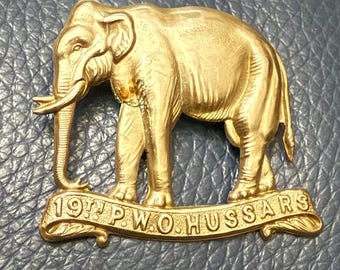 Op de afbeelding: Een gouden metalen badge met een olifantontwerp en de tekst "19th P.W.O. HUSSARS" op een blauwe achtergrond.