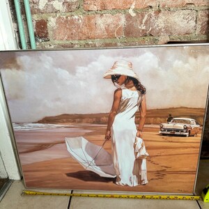 May include: A framed painting of a woman in a white dress and hat standing on a beach with a white umbrella. The woman is looking down at the umbrella. There is a car in the background.