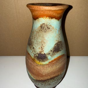 Puede incluir: Un jarrón de cerámica con un esmalte marrón, naranja y azul. El jarrón tiene un cuerpo redondeado y un cuello estrecho.