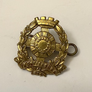 May include: A gold-coloured metal badge with a wreath of leaves surrounding a central design. The design features a cross with a circle in the centre, a crown above the cross, and the words "FRONTIERSMEN" in a curved banner below.