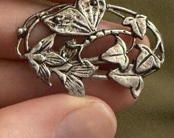 May include: Silver-toned brooch featuring a botanical design. The brooch has an oval shape with a central branch and detailed leaves. The leaves and branch are intricately designed, with a textured finish. The brooch is held in a hand against a green background.
