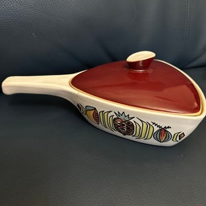 May include: A white ceramic triangular casserole dish with a red lid and a handle. The dish features a colourful fruit design with a pineapple, a strawberry, and other fruits.