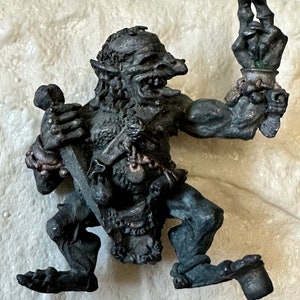 May include: A small, gray, goblin-like figurine with a toothy grin, holding a small club in one hand and a cup in the other. The figurine is made of metal and has a detailed, textured surface.