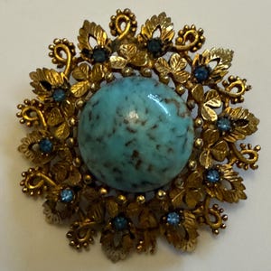 May include: Vintage-style brooch with a large, round, turquoise-colored stone center. The brooch is surrounded by ornate gold-toned metalwork with leaf and floral details, and small blue accent stones. A classic jewelry piece.