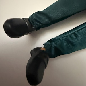 Puede incluir: Primer plano de las piernas de una mu&ntilde;eca. La mu&ntilde;eca lleva pantalones verde oscuro y zapatos negros. Los pantalones son de tela texturizada. Los zapatos son lisos y brillantes.