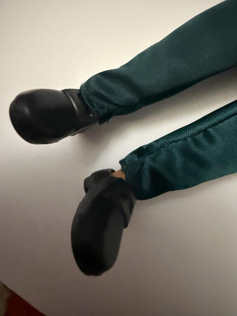 Puede incluir: Primer plano de las piernas de una mu&ntilde;eca. La mu&ntilde;eca lleva pantalones verde oscuro y zapatos negros. Los pantalones son de tela texturizada. Los zapatos son lisos y brillantes.
