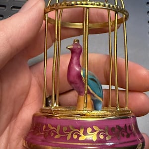 May include: A decorative bird figurine inside a gold-colored cage. The bird has a purple body, blue wings, and a yellow beak. The cage sits on a pink base with gold floral accents.