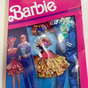 May include: A Barbie Fantasy Fashions set in a pink box. The box features the Barbie logo and images of Barbie and Ken dolls in fantasy outfits. The set includes two complete fashions with blue, red, and gold accents. The text "2 Complete Fashions!" is visible.