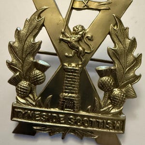 May include: A gold-colored metal badge with a large 'X' in the center. The 'X' is surrounded by a wreath of leaves and thistles. A lion holding a flag with an 'X' on it is in the center of the 'X'. A tower is below the lion. The words 'TYNESIDE SCOTTISH' are on a banner below the tower.