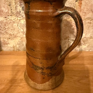 Puede incluir: Una jarra de cerámica marrón con una boca ancha y un asa. La jarra tiene un aspecto rústico y hecho a mano con un esmalte marrón moteado.