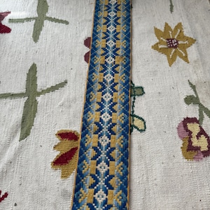 May include: A long, narrow textile with a geometric pattern in shades of blue, yellow, and white. It features a metal clasp and is displayed on a patterned white rug with floral designs.