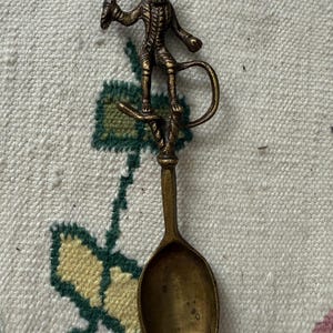 May include: A vintage brass spoon with a monkey figure holding a sword. The monkey is standing on a curved branch above the spoon's bowl. The spoon has a long handle and a rounded bowl.