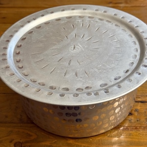 May include: A round, silver metal serving dish with a lid. The lid has a decorative pattern of lines radiating from the center and a ring of small holes around the edge. The sides of the dish have a textured, dotted pattern.