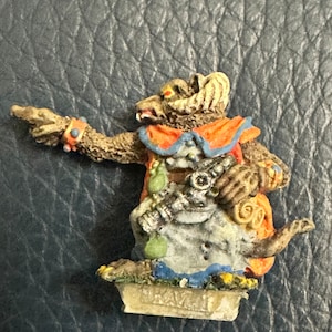 May include: A painted miniature figurine of a furry creature with horns, wearing a red and blue robe, holding a staff and a scroll. The figurine is standing on a small base.