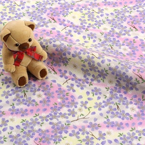 May include: A white fabric with a repeating pattern of purple flowers and green leaves. The fabric has a subtle pink and yellow ombre effect. A brown teddy bear is sitting on the fabric.