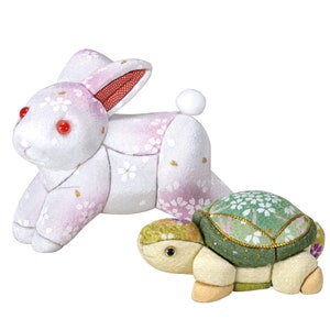 Könnte beinhalten: Ein weißes Kaninchen und eine grüne Schildkröte aus Stoff mit einem Blumenmuster. Das Kaninchen hat rote Augen und befindet sich in einer Laufposition. Die Schildkröte hat einen hellbraunen Kopf und befindet sich in einer Gehposition.